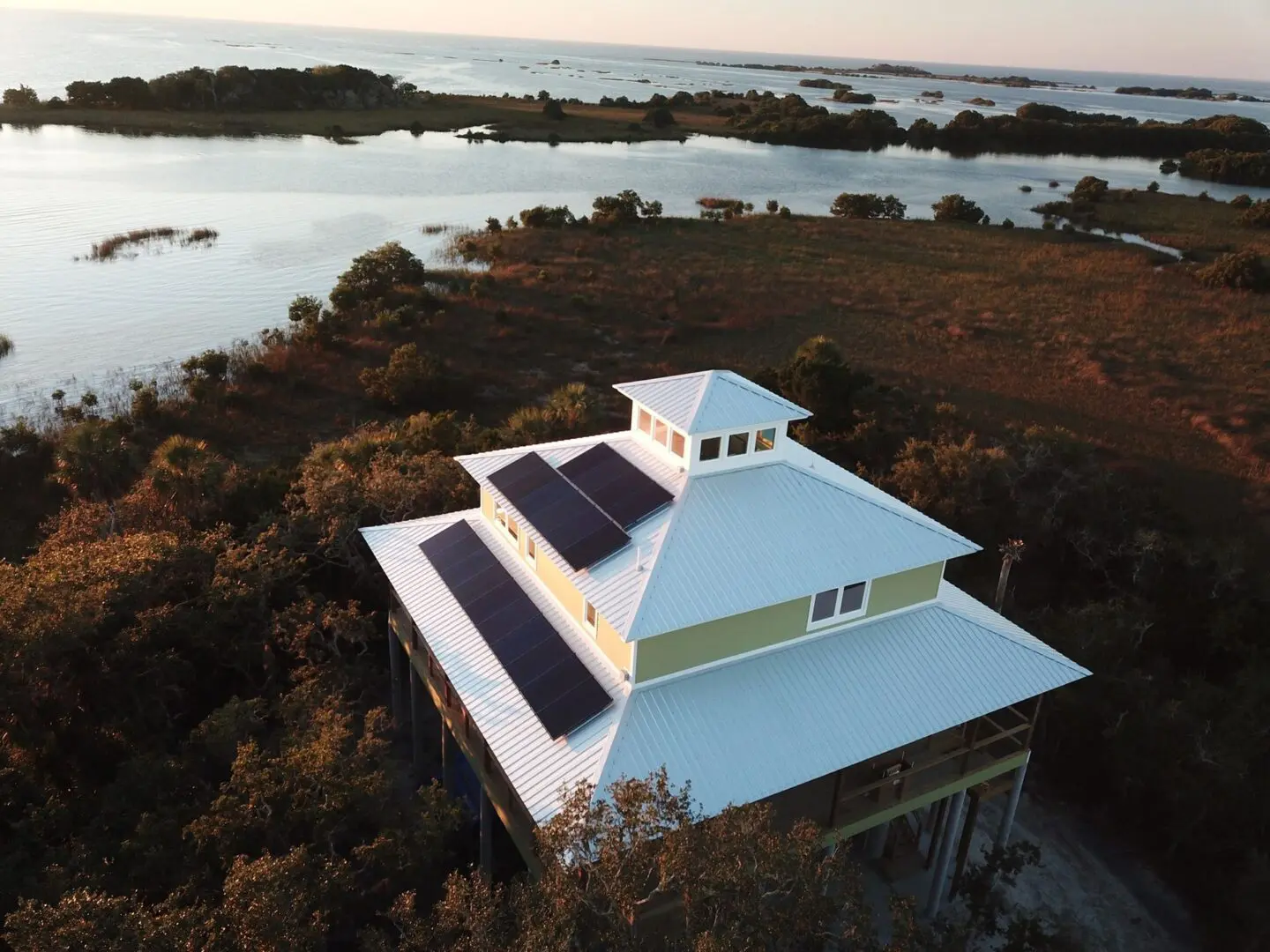 Elevated house with solar panels on a white roof, surrounded by trees and overlooking a coastal water landscape during sunset.