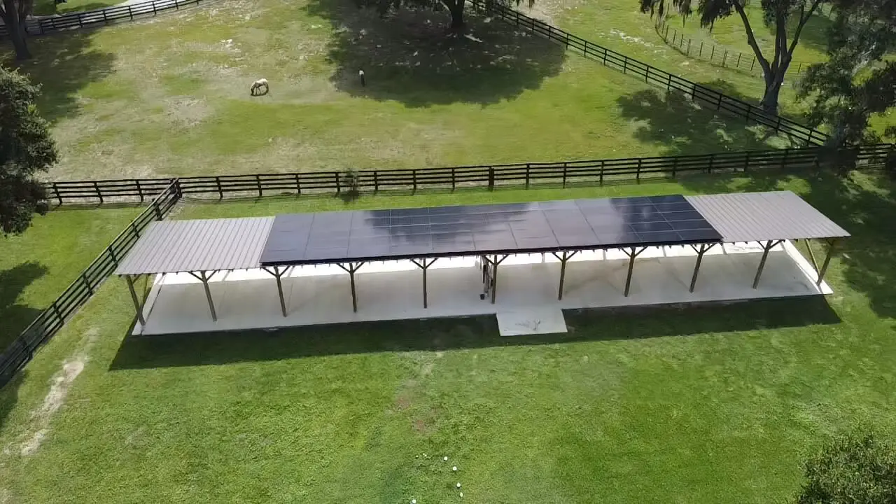Aerial view of a long, covered outdoor structure with solar panels on the roof, situated in a grassy, fenced area with trees and a grazing animal.