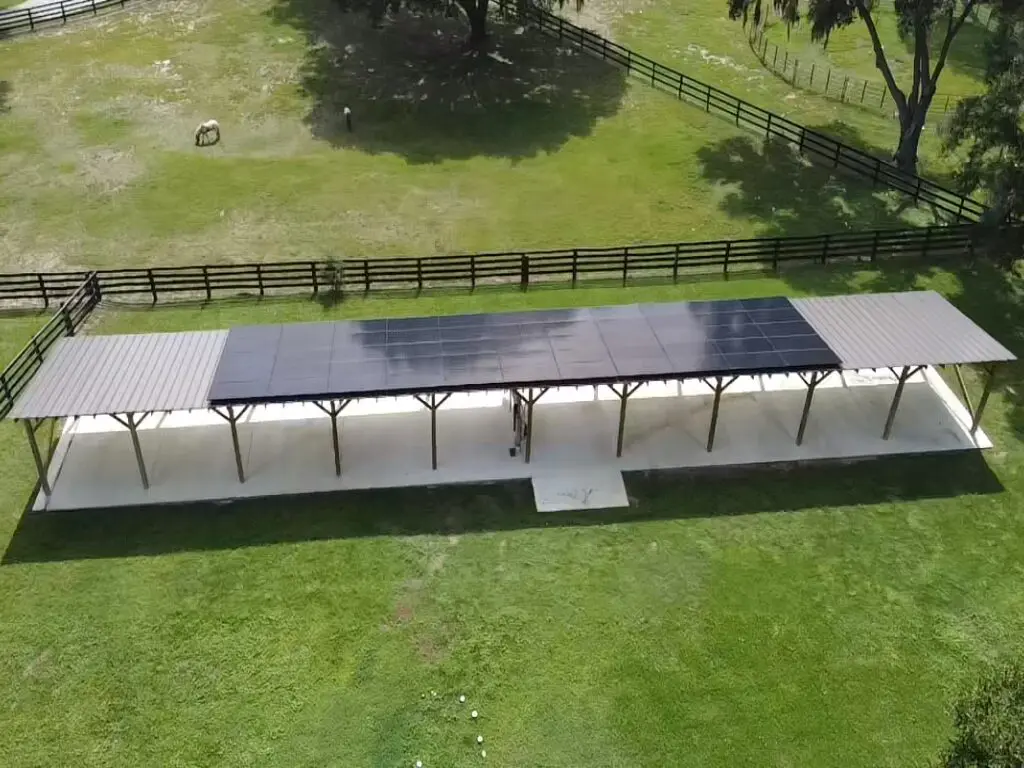 Aerial view of a long solar panel installation on a metal roof structure in a grassy yard with a wooden fence and trees.