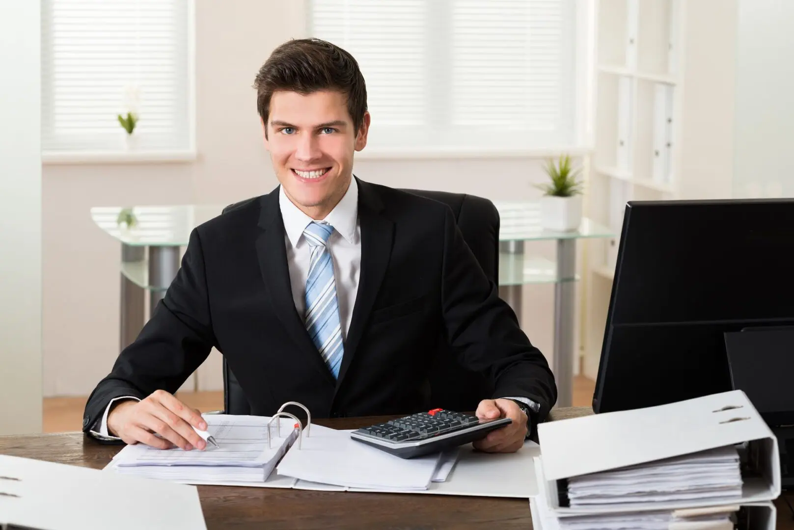 Man in a suit sitting at a desk with documents and a calculator, smiling at the camera.