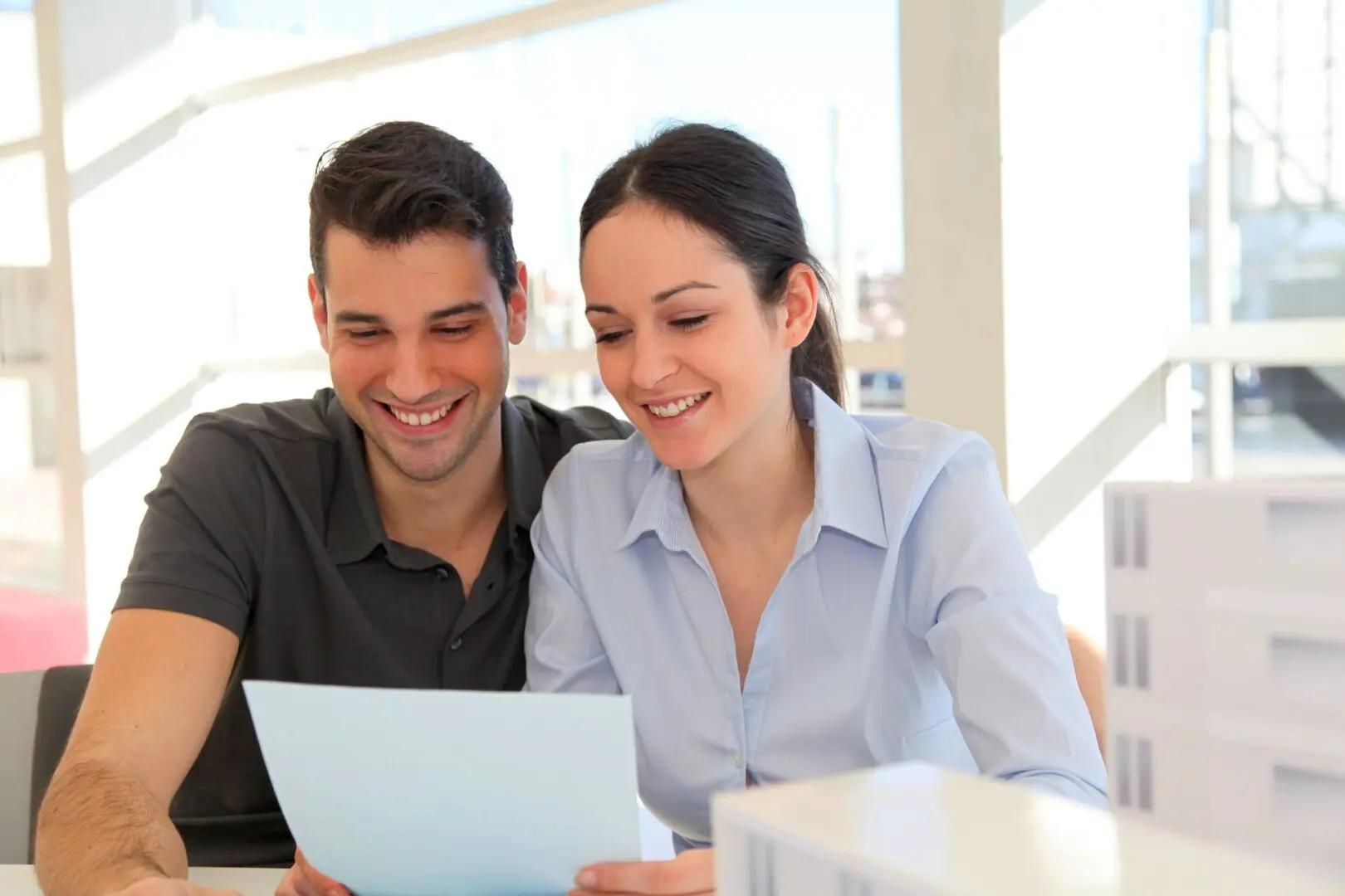 A man and woman sit together at a table, smiling and looking at a piece of paper. They appear to be in a bright, well-lit room.