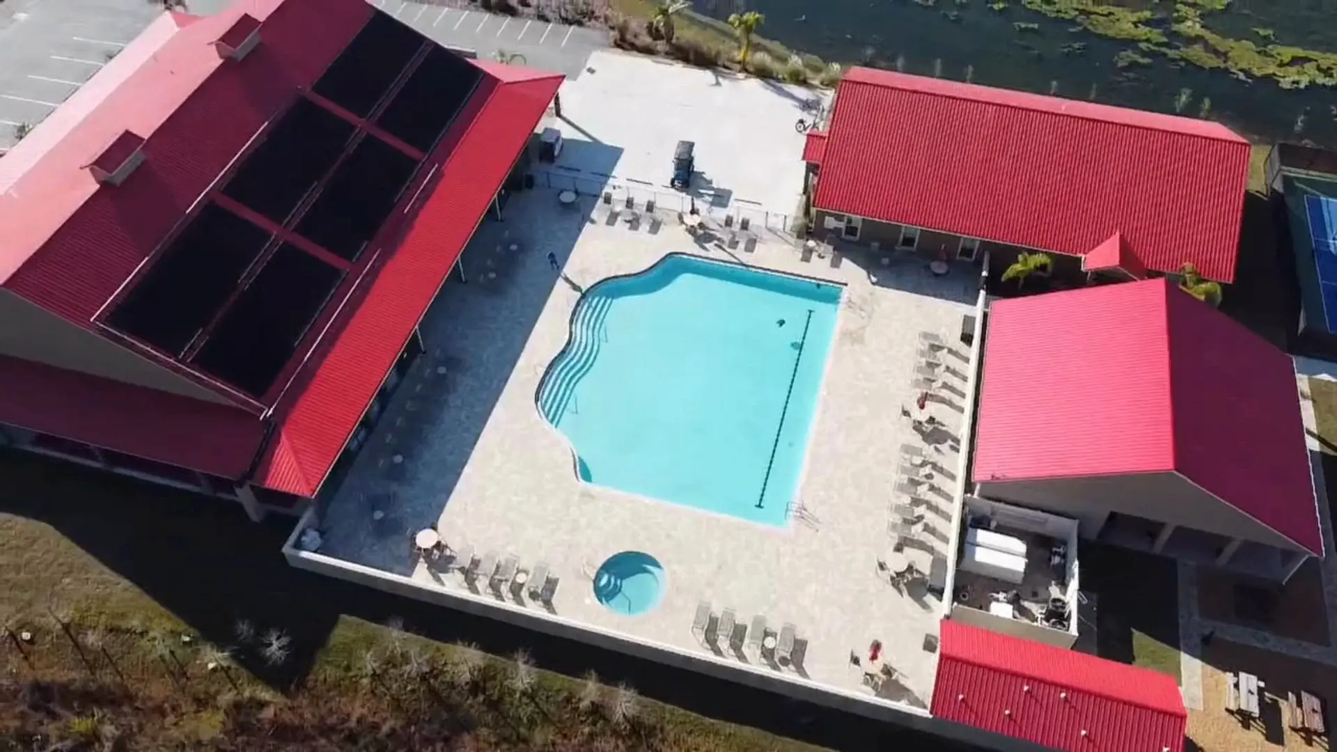 Aerial view of a swimming pool complex with a main pool, a smaller circular pool, red-roofed buildings, and surrounding lounge chairs, adjacent to a parking area and a body of water.