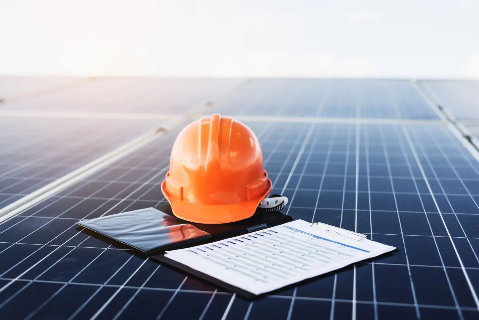An orange hard hat and clipboard with papers rest on a solar panel under a clear sky.