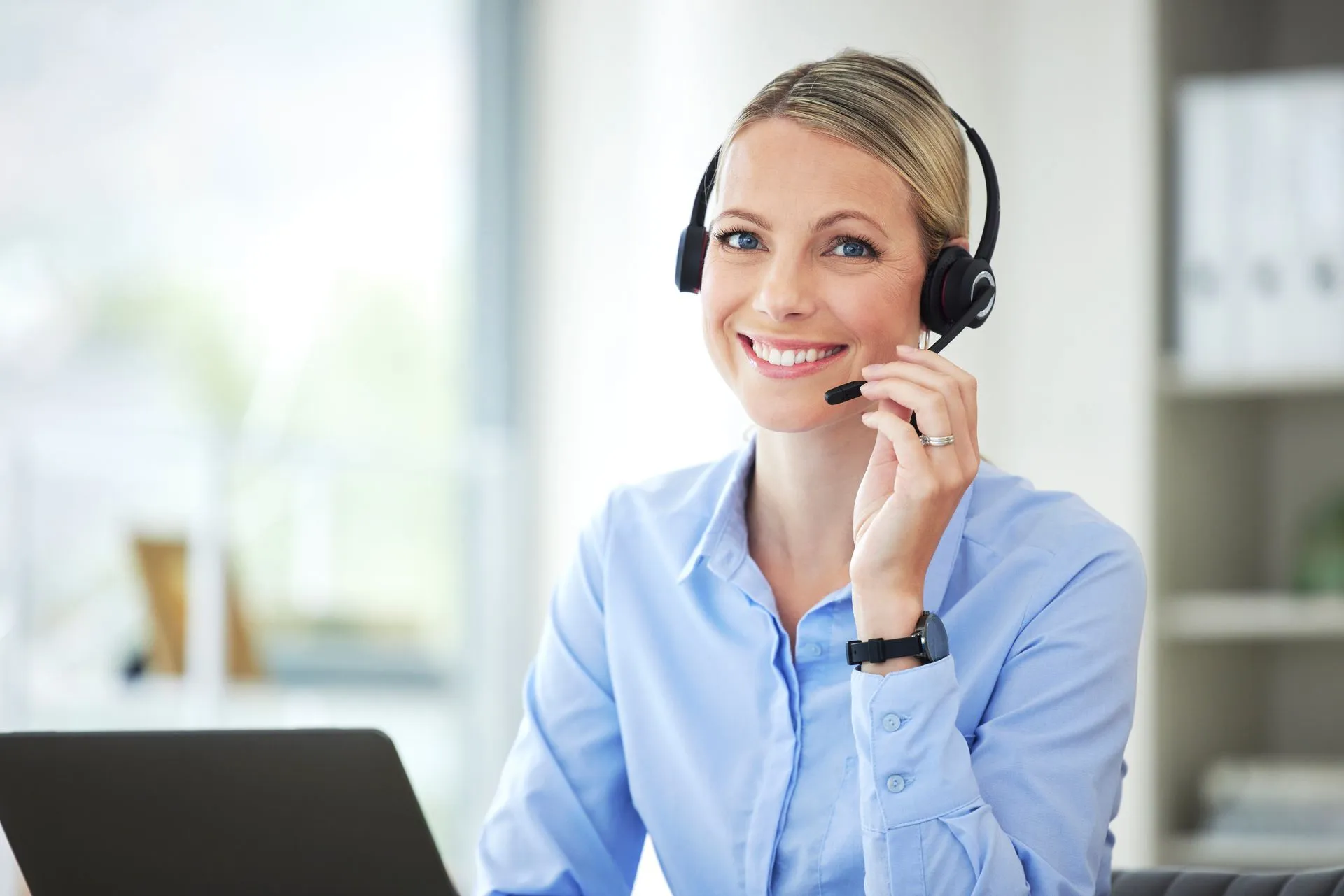 A woman in a blue shirt is sitting at a desk, wearing a headset, and smiling at the camera. She has a laptop in front of her.