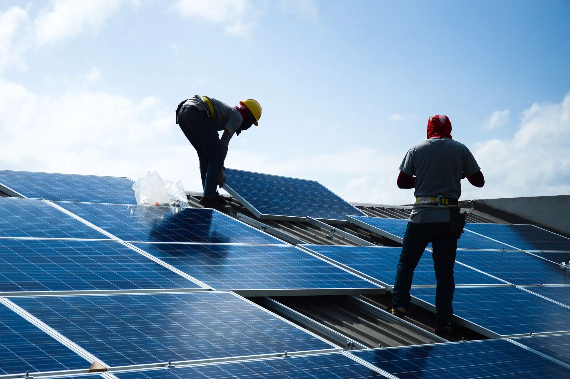 Two workers in safety gear are installing solar panels on a rooftop under a partly cloudy sky.