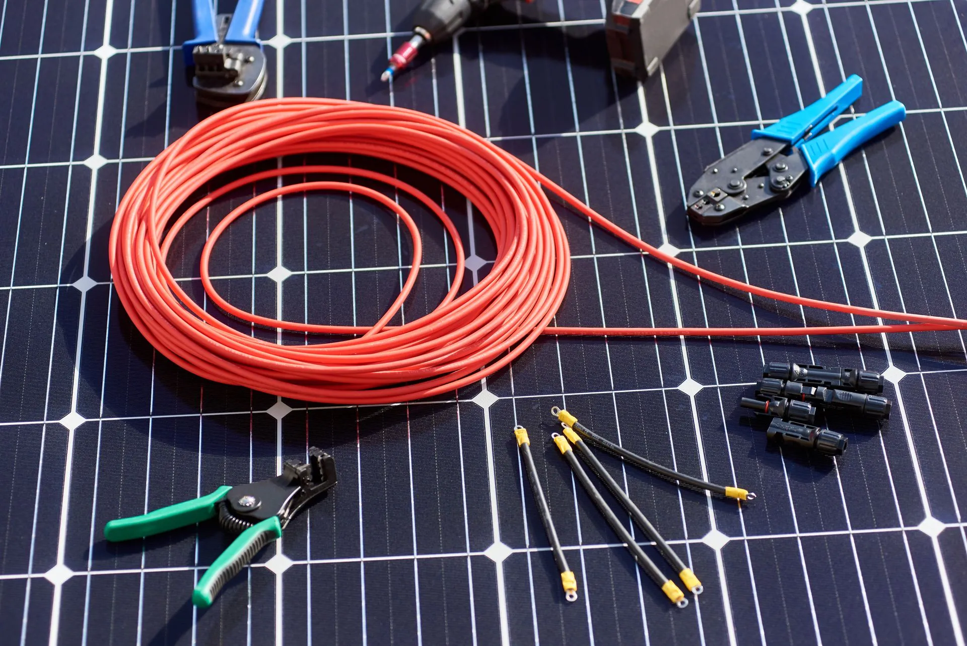 Red electrical cable and various tools, including wire cutters and connectors, are laid out on a solar panel surface.
