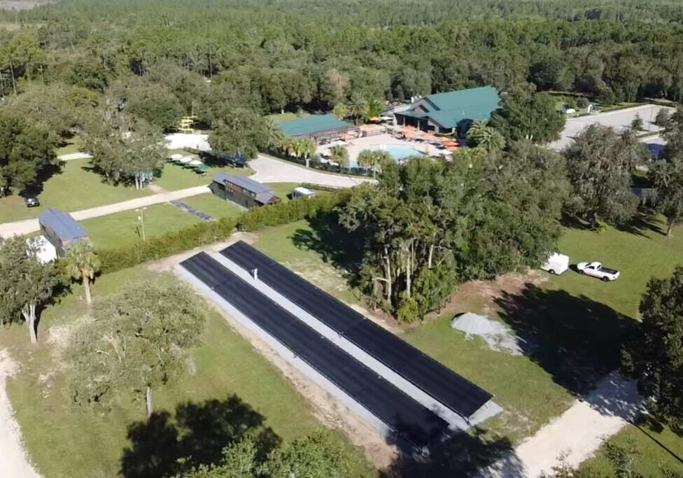 Aerial view of a rural property featuring a building with a green roof, a solar panel array, trees, and surrounding open land.