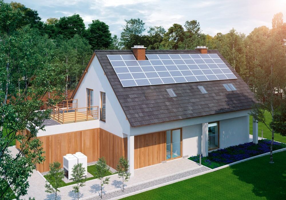 A modern house with solar panels on the roof, set in a green, wooded area. The building features a rooftop terrace and large windows.