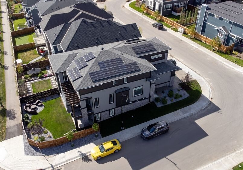 Aerial view of a suburban neighborhood with modern homes. A house with solar panels is in the foreground. Two cars, one yellow and one black, are parked next to it on the street.