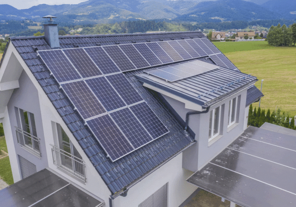 A modern house with solar panels on a sloped roof, surrounded by green fields and distant mountains under a cloudy sky.