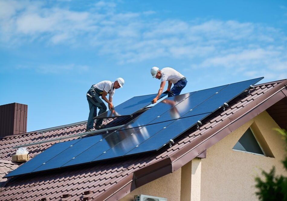Two workers in helmets install solar panels on a sloped house roof under a clear blue sky.