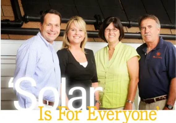 Four people stand smiling in front of a solar panel with the text "Solar Is For Everyone" in the foreground.