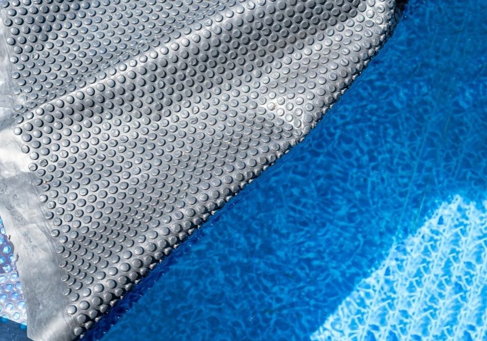 Close-up of a partially unrolled silver bubble wrap pool cover next to clear blue water, casting a shadow on the bottom of the pool.