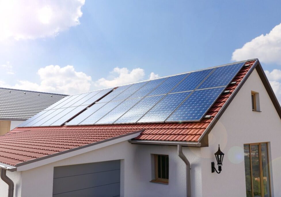 Solar panels installed on the red-tiled roof of a white house with blue sky and clouds in the background.