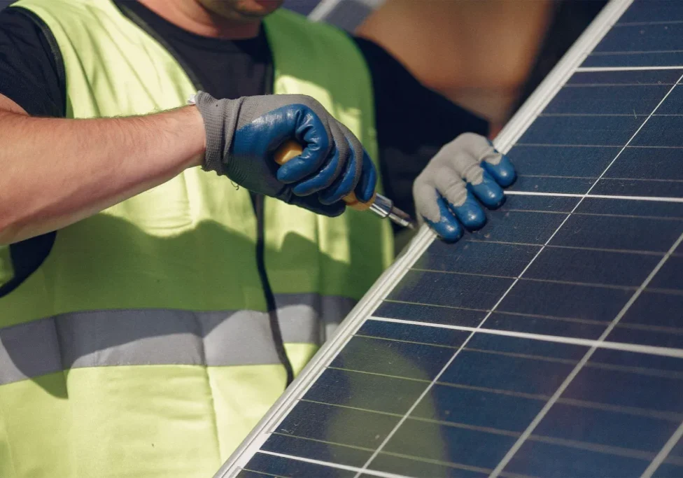 Worker in a reflective vest and gloves using a screwdriver to install a solar panel.