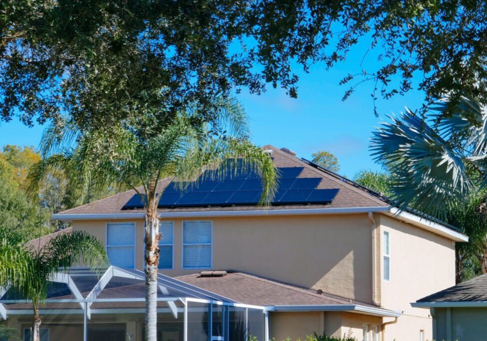 House with solar panels on the roof, surrounded by palm trees and a screened porch, under a clear blue sky.