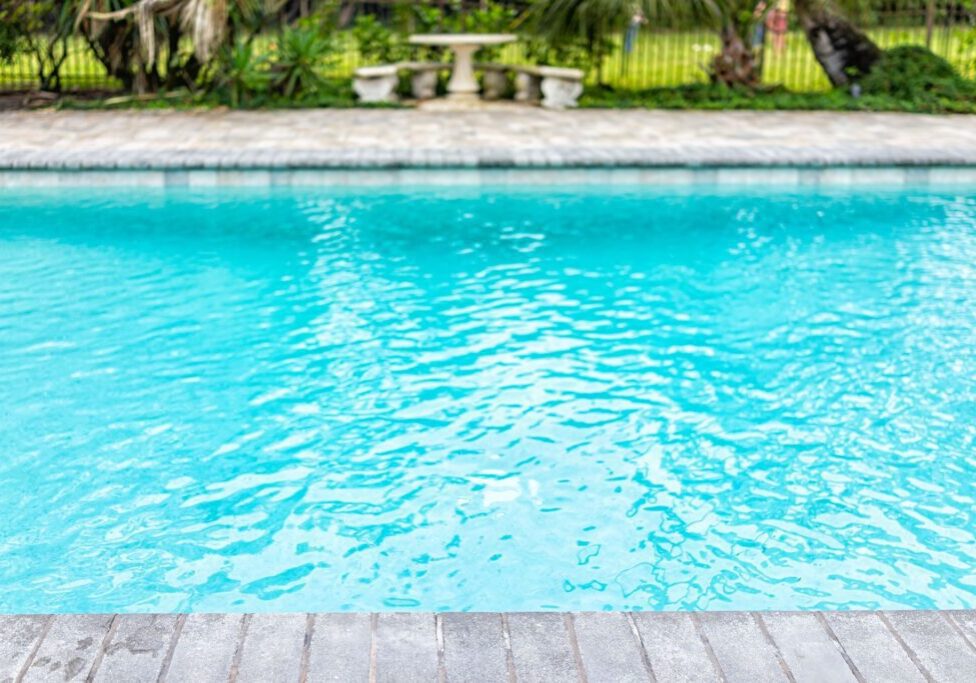 A clear blue outdoor swimming pool with a stone deck. In the background, there are trees, plants, and stone benches near a small table.
