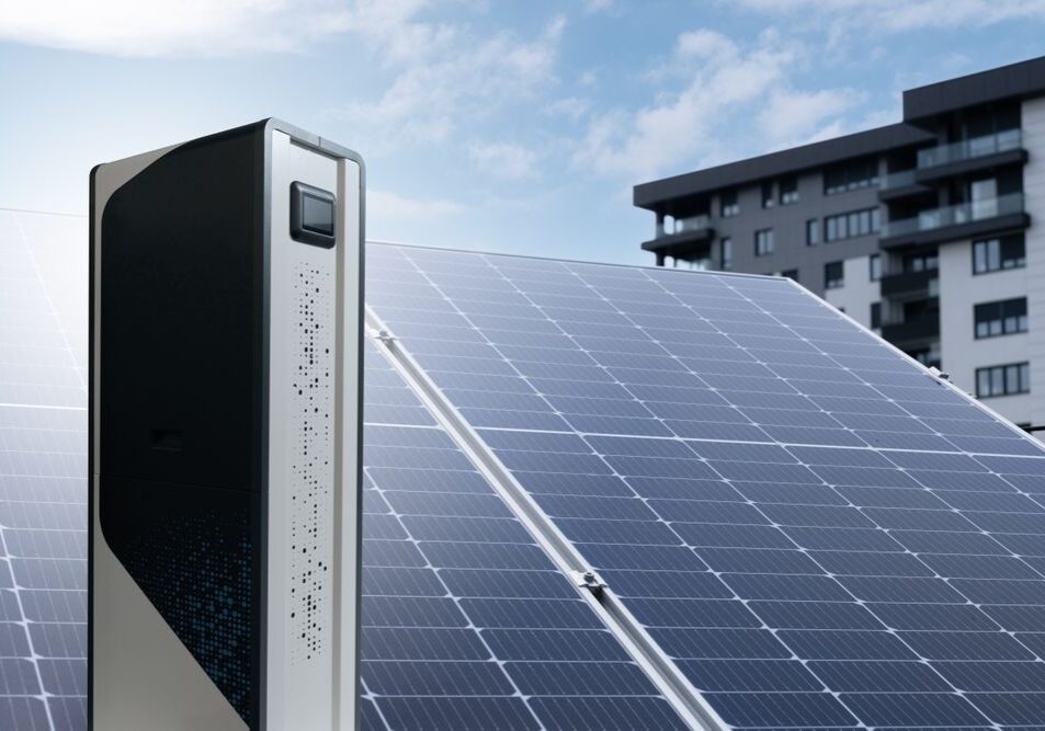 A solar panel array with a modern battery storage unit is set against a clear sky, with a multi-story residential building in the background.