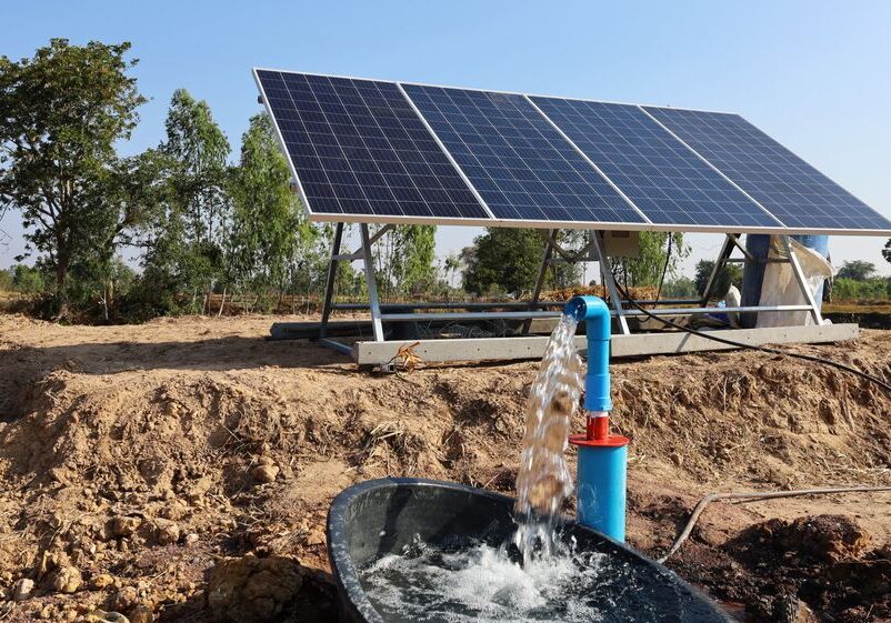 Solar panels power a pump, drawing water into a black trough in a rural setting under a clear blue sky.
